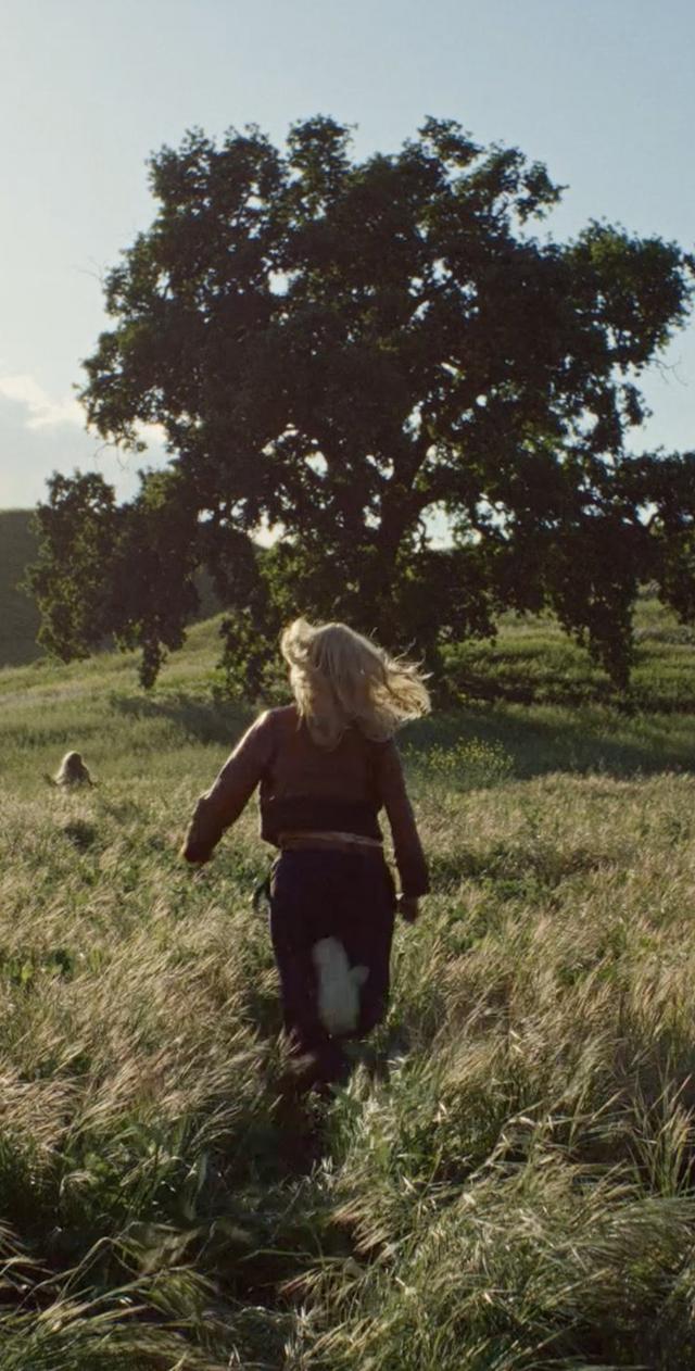 a woman walking through a field with a tree in the background