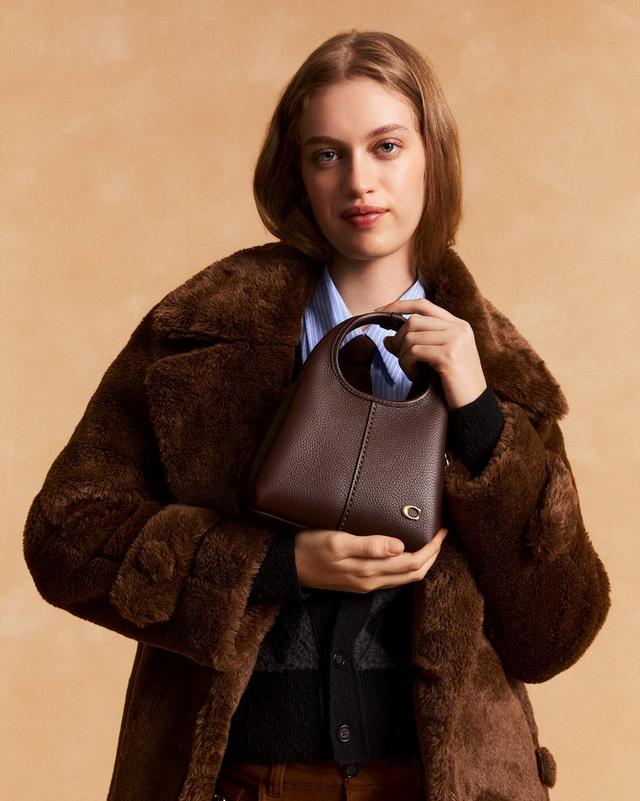 Model holding a brown leather handbag with circular handle, wearing a brown faux fur coat.