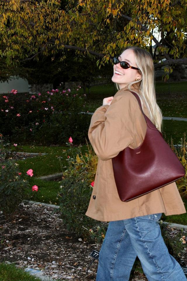 Woman wearing sunglasses and tan coat carrying burgundy leather shoulder bag in a garden setting.