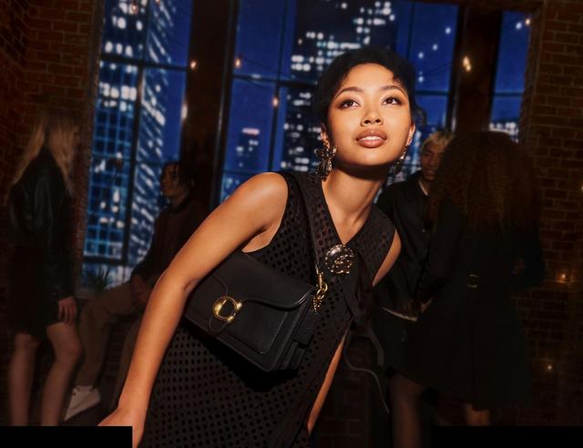 Woman wearing black textured dress with statement earrings and black crossbody bag at evening event.