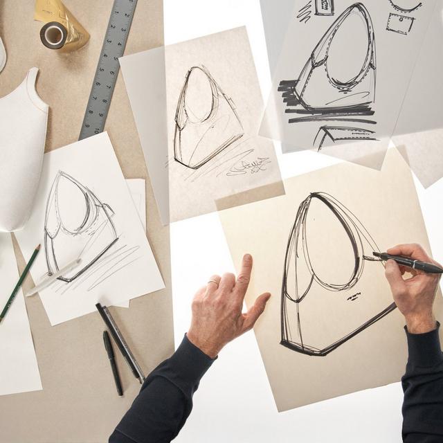 Designer sketching handbag concepts with pen on paper surrounded by design tools and reference sketches.