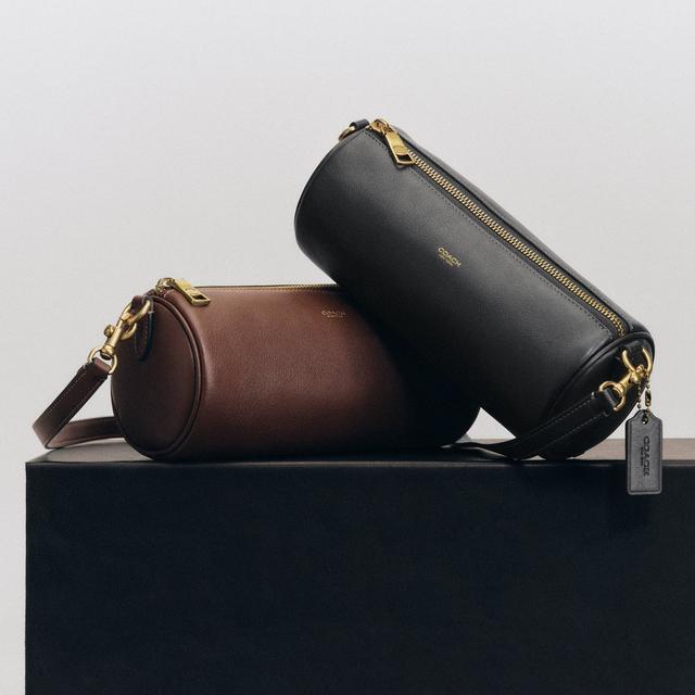 Two Coach leather crossbody bags with gold hardware, one brown and one black, displayed on a box.