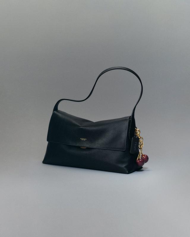 Coach black leather shoulder bag with fold-over flap and cherry charm attached to gold hardware.