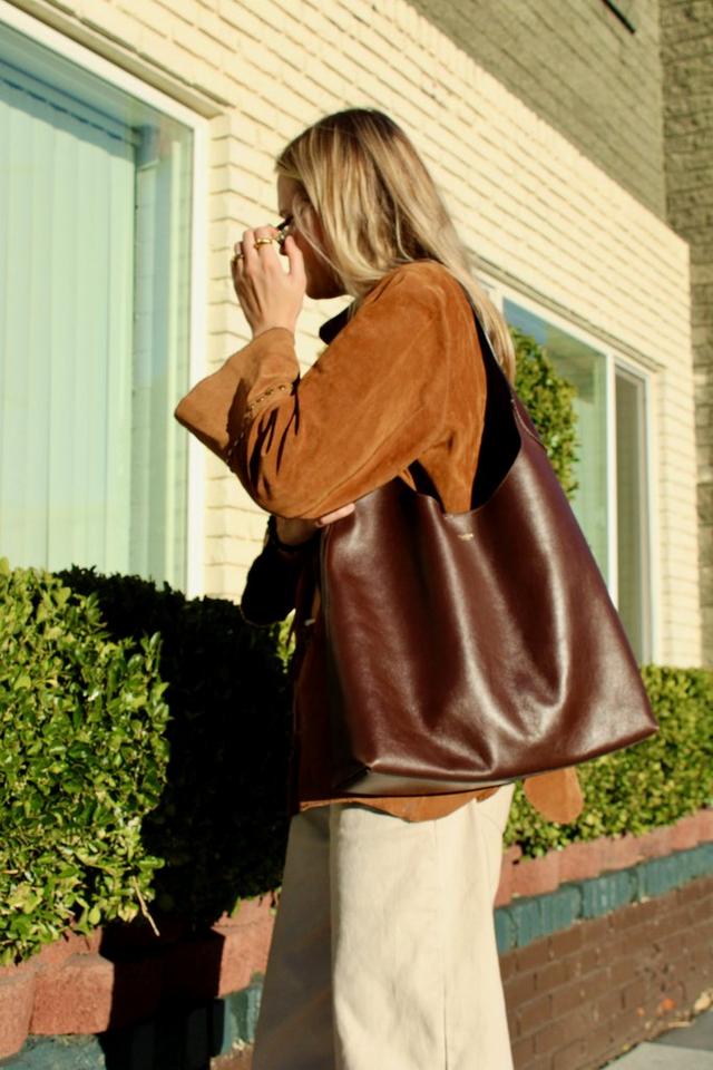 Woman wearing brown suede jacket and white pants carrying large brown leather tote bag on her shoulder.