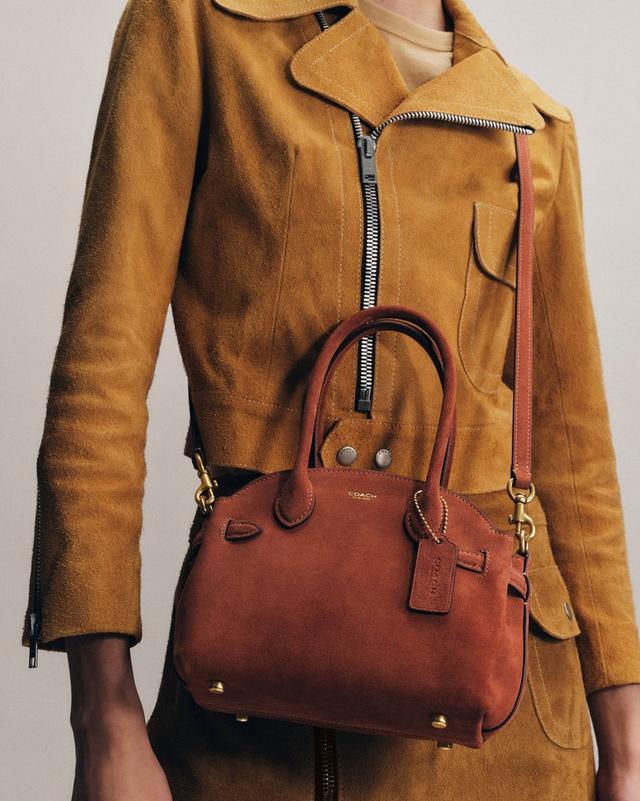 Person wearing tan suede jacket with Coach brown leather crossbody bag with top handles and detachable strap.