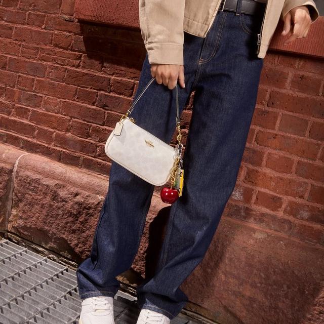 Person wearing beige jacket, red top, dark jeans, and white sneakers, holding a white Coach crossbody bag.