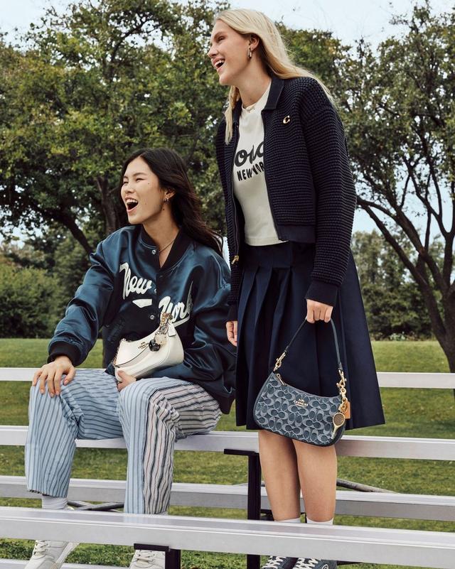 Two women laughing together outdoors, wearing Coach jackets and carrying Coach handbags.