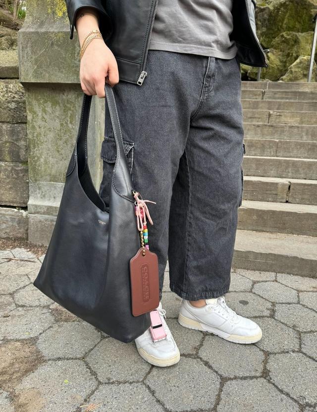 Person holding black leather Coach shoulder bag with colorful charm and brown leather tag attachment.