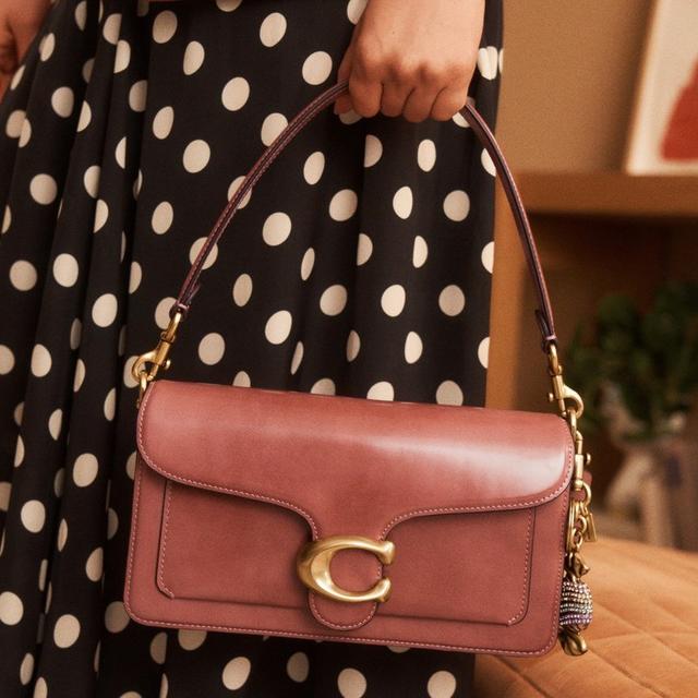 Person holding a brown leather shoulder bag with gold hardware, wearing a black polka dot dress.