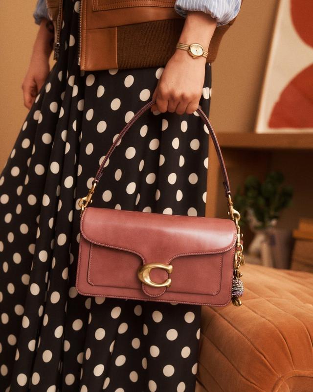 Woman holding a brown leather shoulder bag with gold hardware, wearing a polka dot skirt and gold watch.