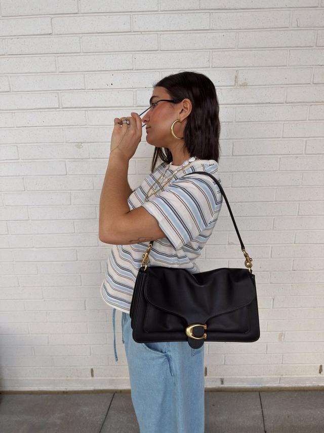 Woman wearing black leather shoulder bag with gold hardware, styled with striped shirt and jeans.