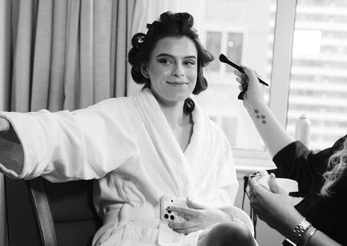 Woman in white robe having makeup applied while wearing hair rollers and holding phone.