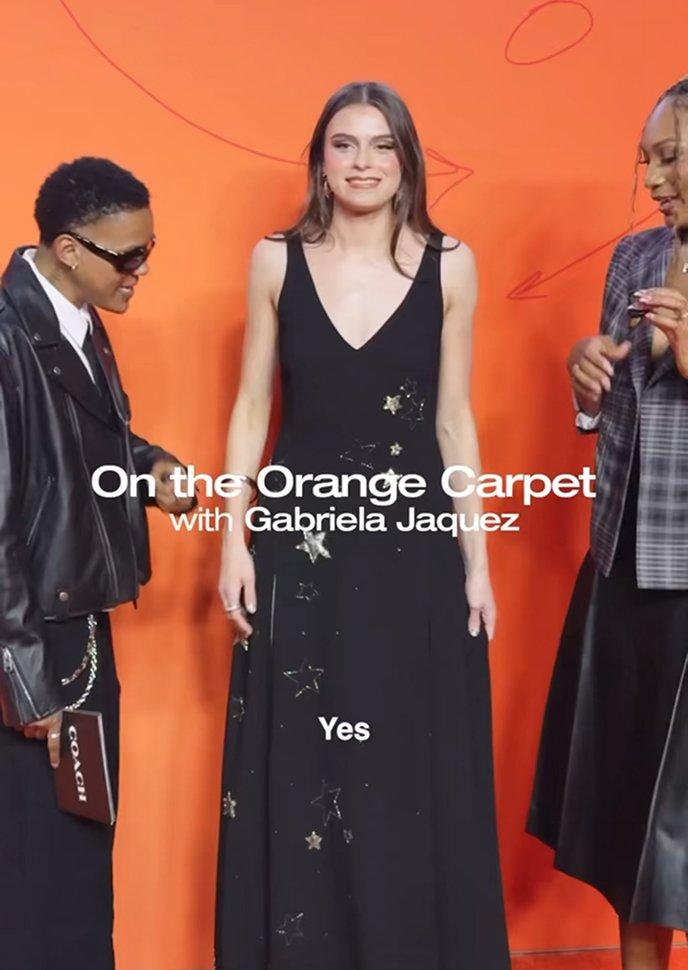 Woman in black sleeveless dress with star embellishments posing on orange carpet at event.