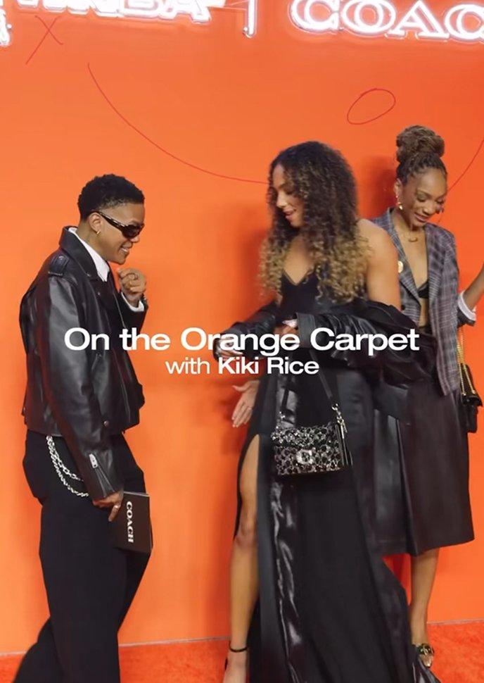 Three people posing and interacting on an orange carpet at an event.
