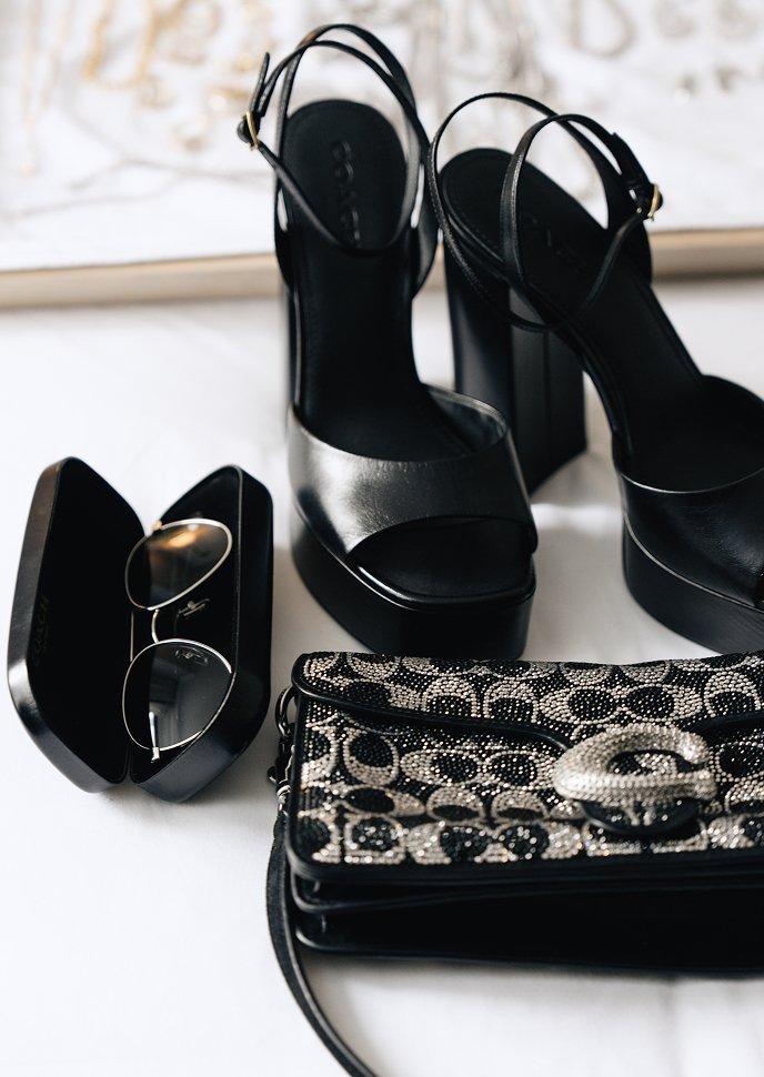 Black strappy platform heels, sunglasses, and an embellished clutch arranged together.