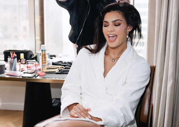 Woman in white robe laughing while getting her hair styled, with makeup products visible on table.