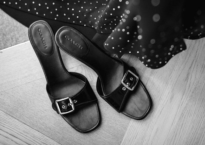 Coach black leather slide sandals with silver buckle detail on wooden floor beside sequined fabric.