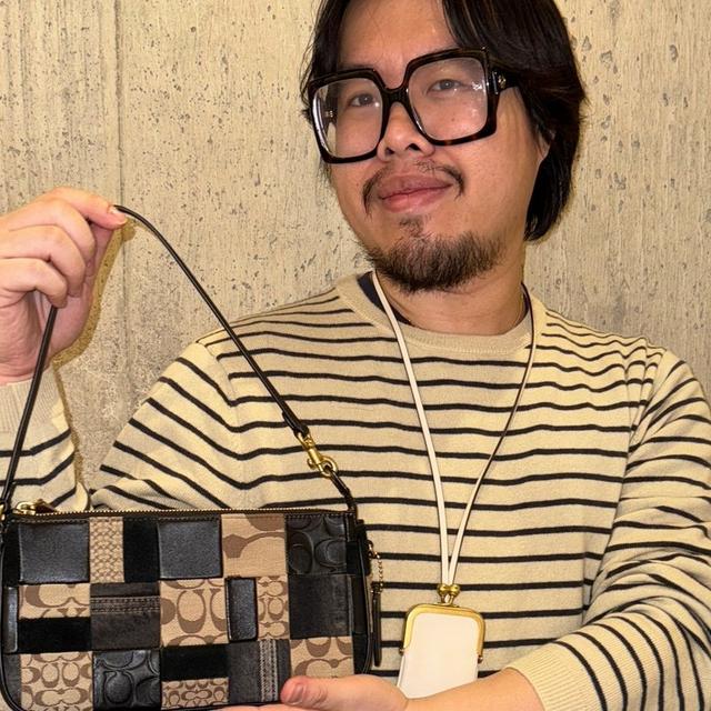 Person wearing black-framed glasses and striped shirt displaying a Coach patchwork shoulder bag with strap.