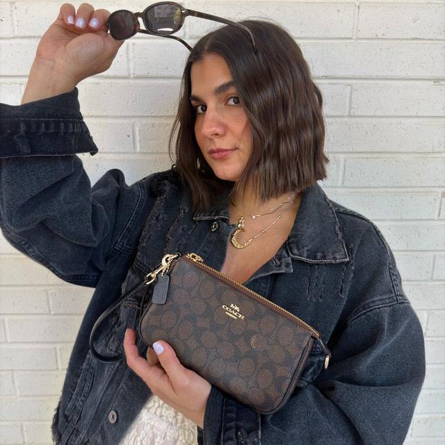 Woman wearing denim jacket holding sunglasses and Coach crossbody bag with layered gold necklaces.