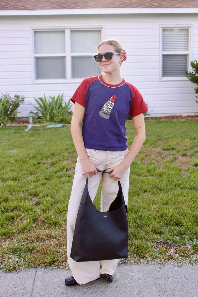 Person wearing sunglasses and a purple raglan tee with red sleeves, holding a black tote bag outdoors.