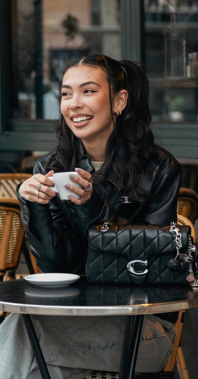 Woman in black leather jacket smiling while holding a coffee cup at an outdoor café table with black handbag.