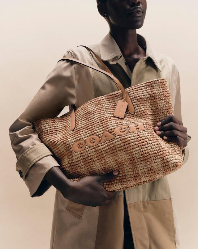 Model wearing beige jacket holding a woven straw Coach tote bag with leather handles and logo.