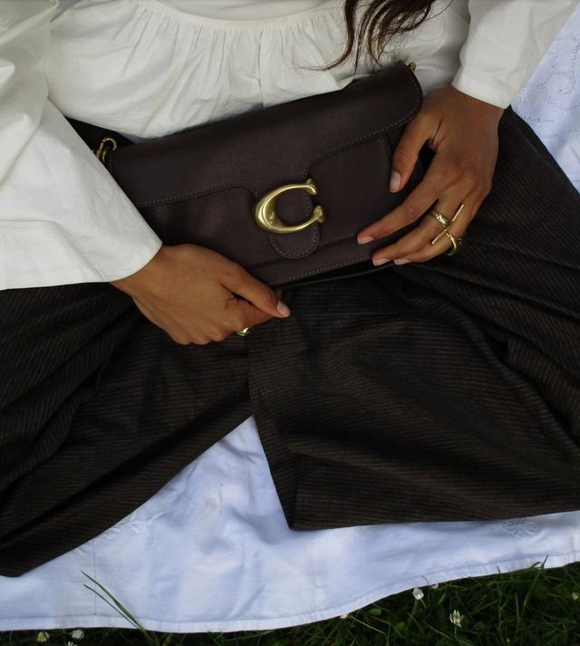 Person wearing white blouse and black pants holding dark brown leather handbag with gold C-shaped clasp.