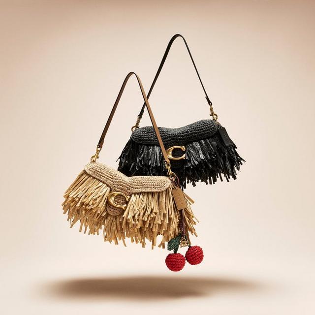 Two woven shoulder bags with fringe detailing, one in beige with cherry charm, one in black.