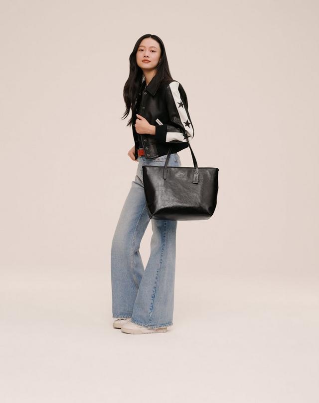 Model wearing black leather jacket with white star-print sleeves, blue wide-leg jeans, and holding a black tote bag.