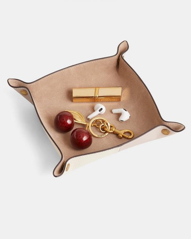 Tan leather valet tray holding a gold lipstick, cherry keychain, wireless earbuds, and small coin.