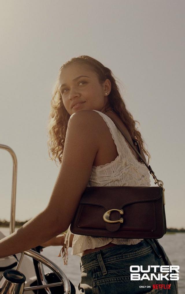Woman wearing white eyelet top with brown leather crossbody bag riding bicycle near water.