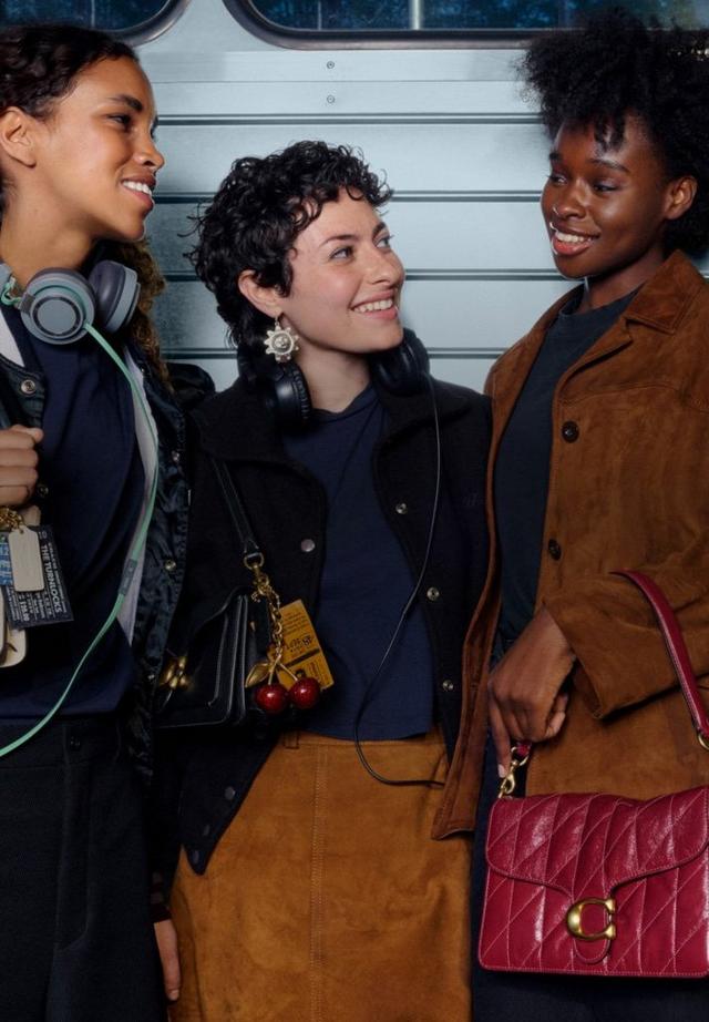 Three women smiling and chatting while wearing jackets and carrying shoulder bags with headphones.