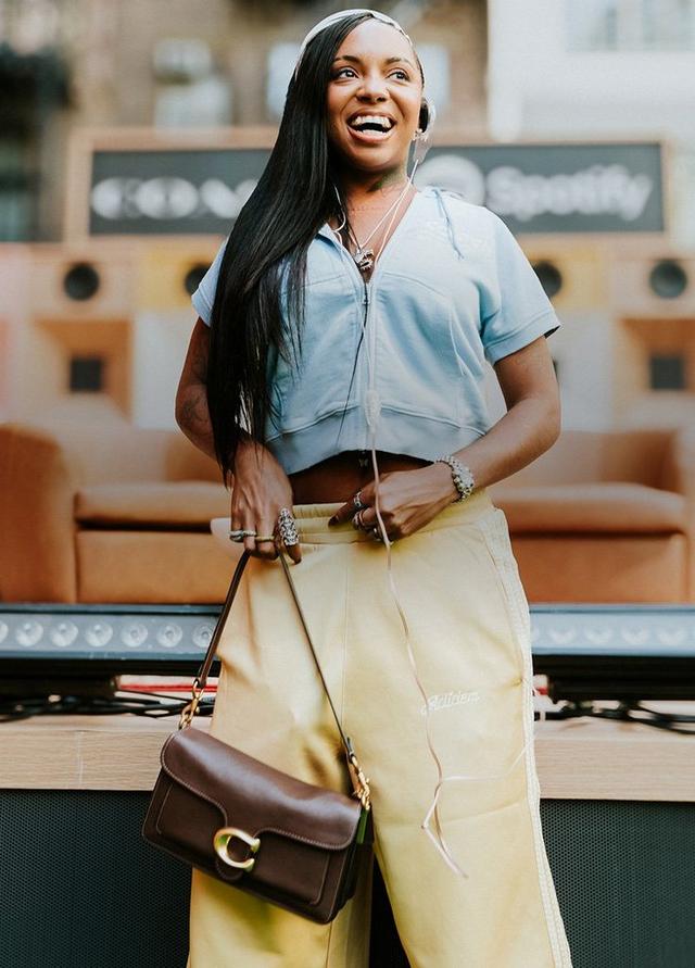 Woman smiling in blue top and yellow skirt holding a brown leather handbag in a modern retail setting.
