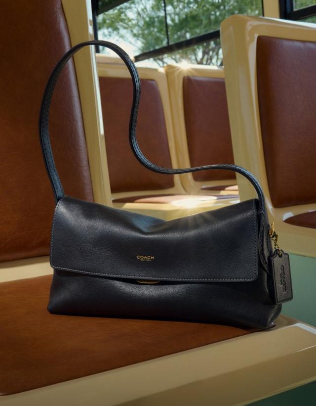 Brown leather Coach shoulder bag with adjustable strap and logo tag displayed on a vintage chair.
