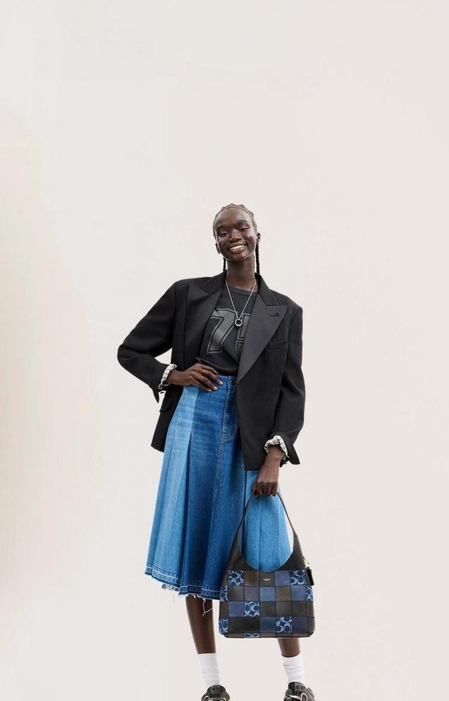 Model wearing black blazer, blue denim skirt, and holding a patterned blue and black shoulder bag.