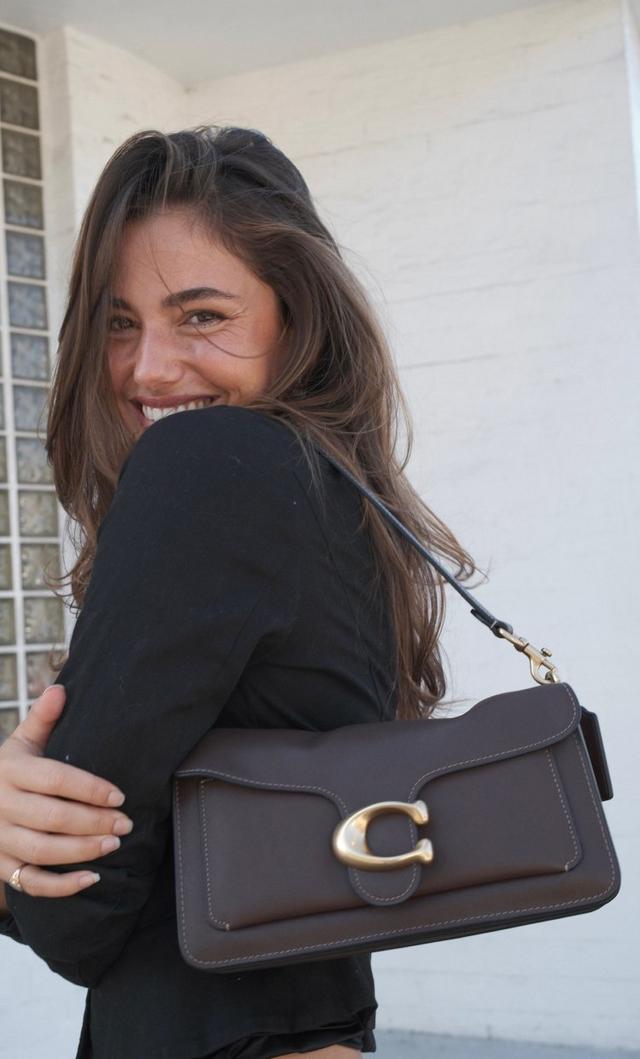 Woman wearing black top smiling while carrying a brown leather shoulder bag with gold hardware clasp.