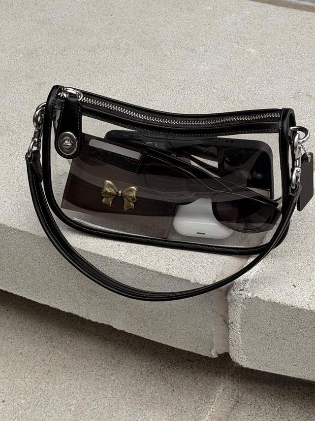 Clear crossbody bag with black trim containing sunglasses, small purse with bow detail, and phone.