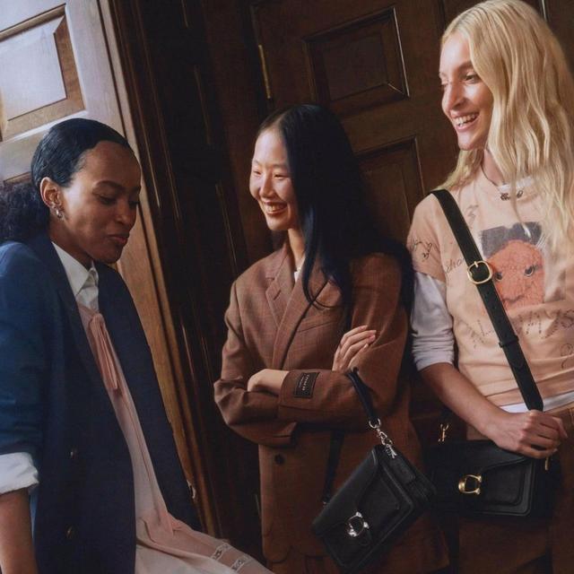 Three women smiling and conversing indoors, each carrying different style shoulder bags with gold hardware.