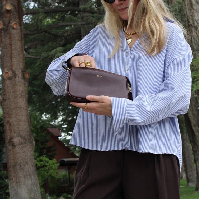 Woman wearing sunglasses and striped shirt holding a Coach brown leather crossbody bag outdoors.