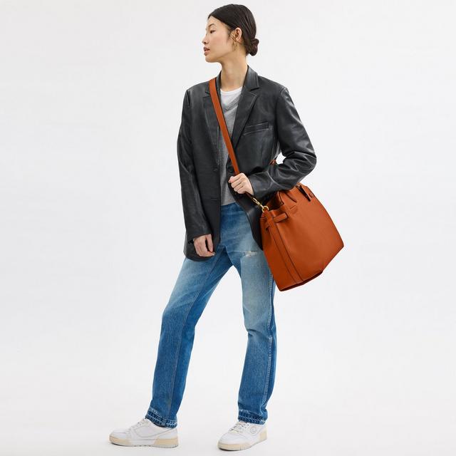 Woman wearing black leather blazer, gray sweater, and blue jeans, carrying an orange leather bucket bag.