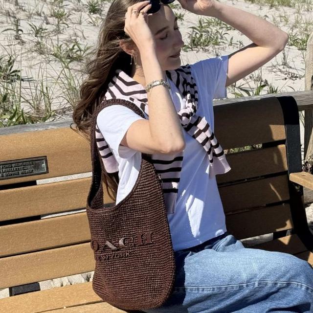 Woman sitting on a bench tying a black scrunchie in her hair while wearing a striped scarf and brown tote bag.