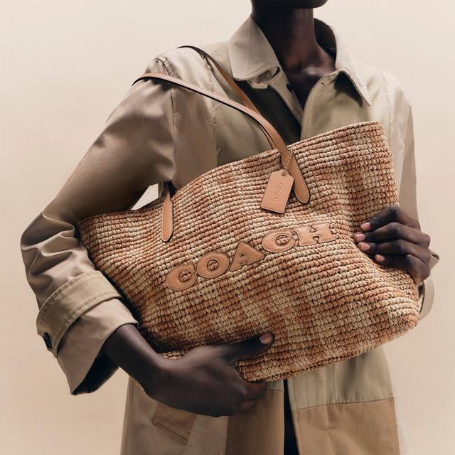 Model holding a woven Coach tote bag with leather handles and logo detail while wearing a beige jacket.