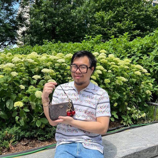 Person wearing glasses holding a brown wristlet with cherry charm keychain outdoors near flowering shrubs.