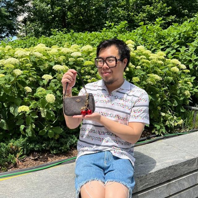 Person sitting on park bench holding small brown wristlet with cherry charm, wearing floral striped shirt.