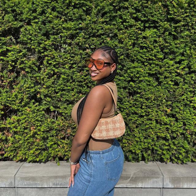 Woman wearing orange-tinted sunglasses, tan crop top, and denim skirt with woven crossbody bag, smiling.