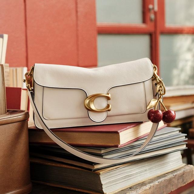 White leather shoulder bag with gold C clasp and cherry charm, displayed on stacked books.