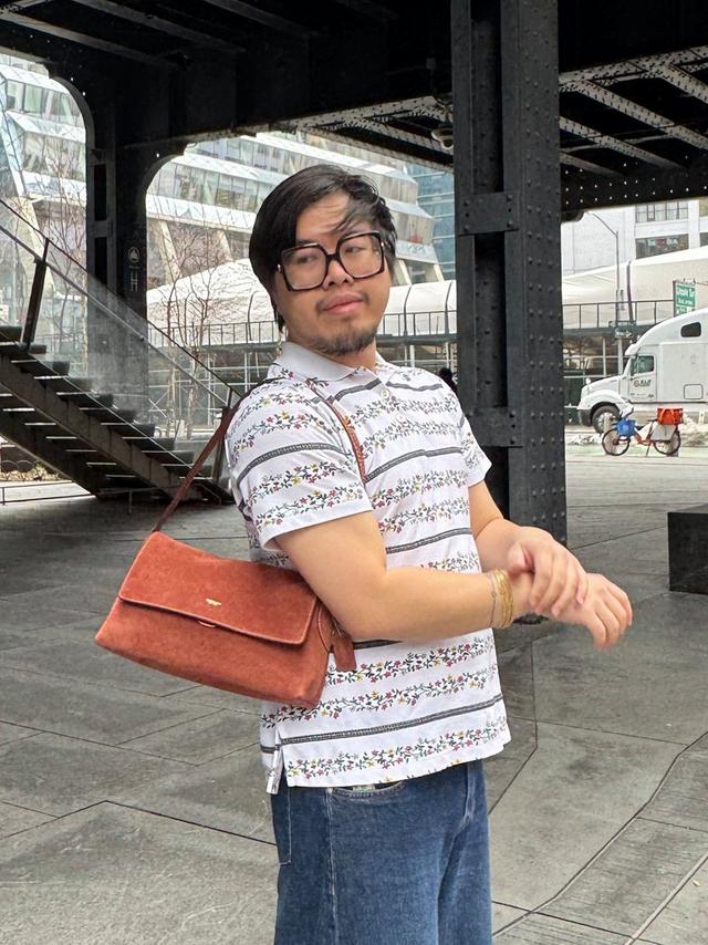 Person wearing large black glasses and white floral striped shirt, carrying a brown suede crossbody bag.