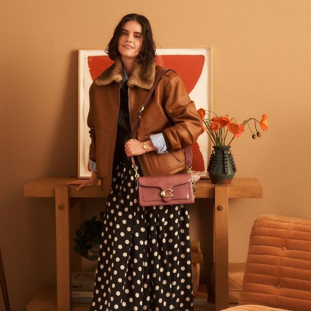 Woman wearing brown leather jacket with fur collar, black polka dot maxi skirt, and pink crossbody bag.