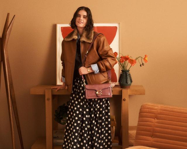 Woman wearing brown leather jacket with fur collar, polka dot skirt, and pink crossbody bag.