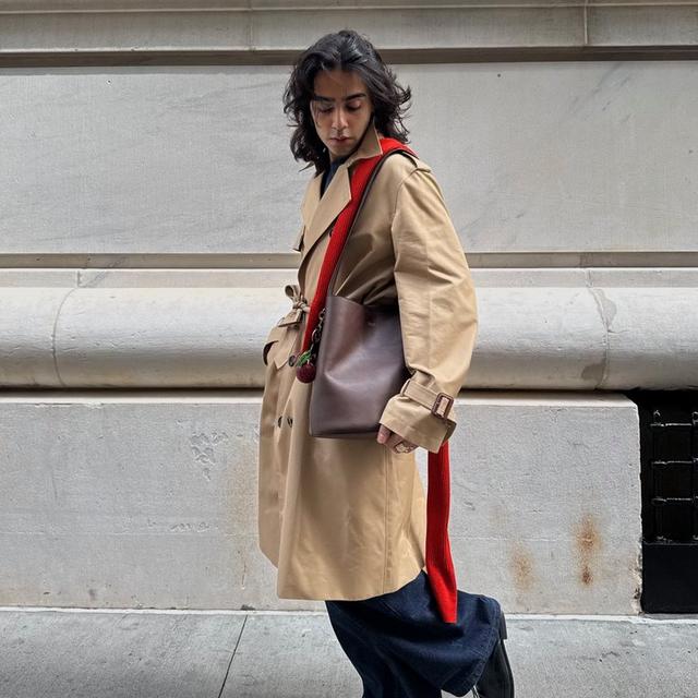 Model wearing tan trench coat with brown leather crossbody bag, red strap, and black wide-leg pants.